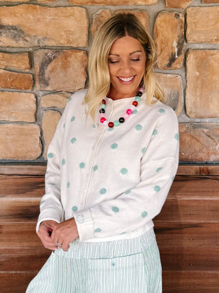 A woman wearing a cream zip-up cardigan with mint green polka dots and a striped skirt, standing in front of a rustic stone wall.