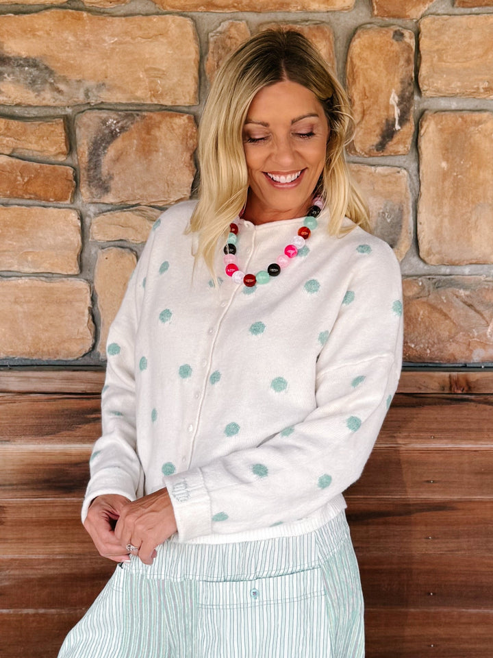 A woman wearing a cream zip-up cardigan with mint green polka dots and a striped skirt, standing in front of a rustic stone wall.