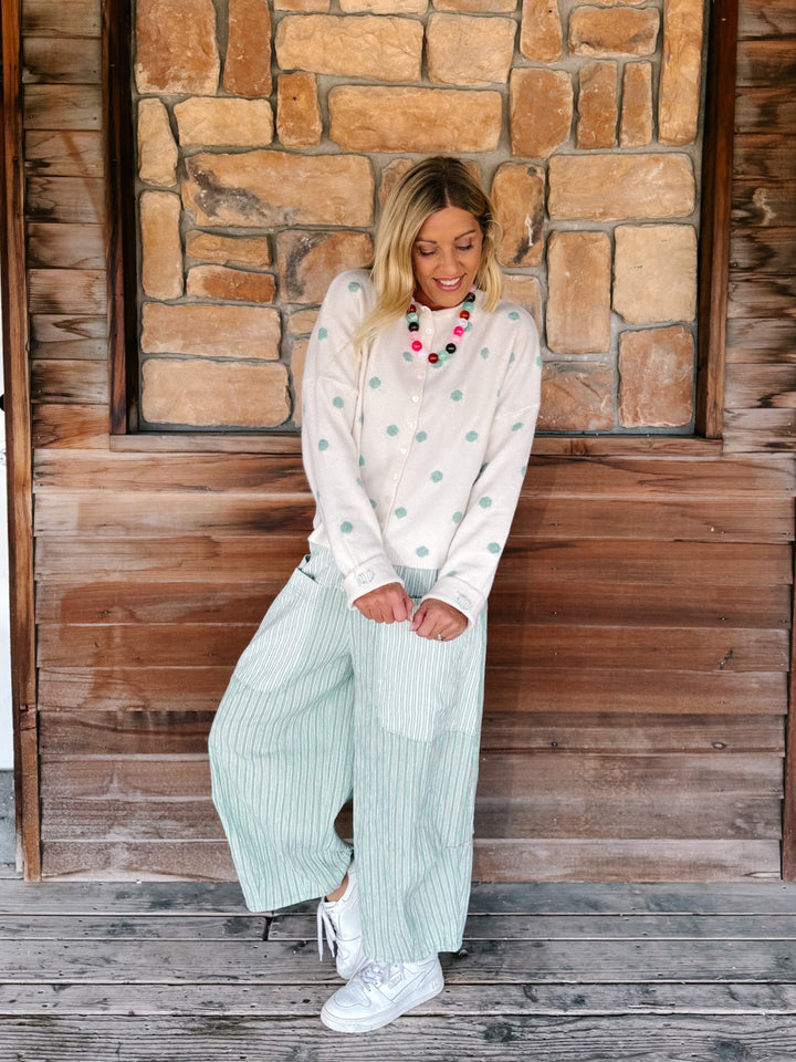 A woman stands on a wooden porch in front of a stone wall, wearing a cream long-sleeve top with mint green polka dots and matching wide-leg mint green striped pants, paired with white sneakers and a colorful beaded necklace.