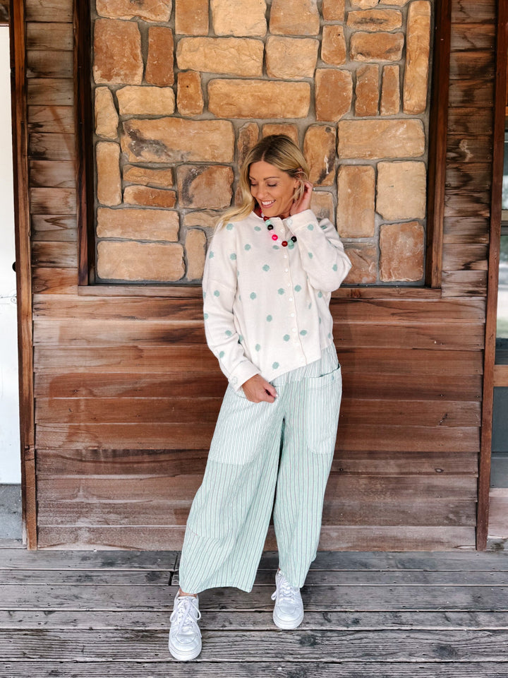 A woman stands on a rustic wooden porch in front of a stone wall, wearing a cream long-sleeve button-up shirt with mint green dots, mint green and white vertically striped wide-leg pants, and white sneakers.