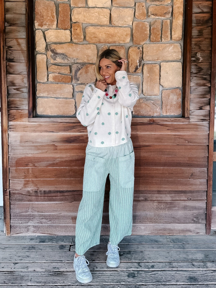 A woman stands on a wooden deck in front of a rustic stone and wood wall, wearing a white long-sleeve button-up shirt with mint green buttons, light green pinstriped wide-leg pants, and white sneakers.