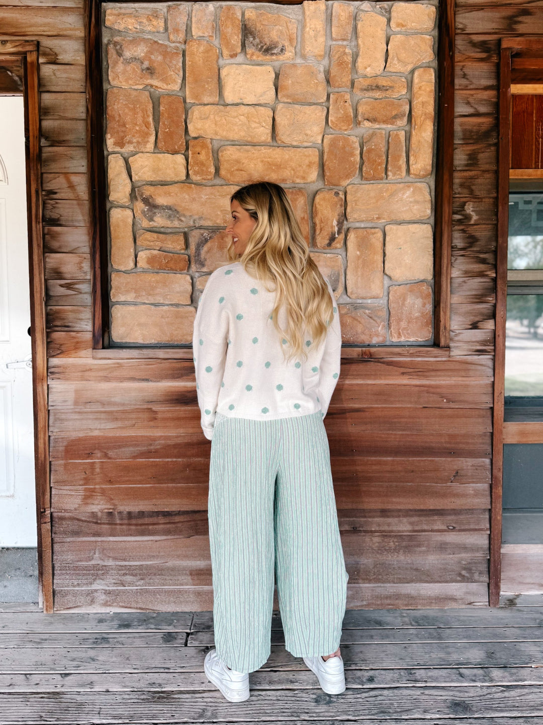 A woman stands on a rustic wooden porch facing a stone wall, wearing a cream knit sweater with mint green polka dots and matching wide-leg ribbed pants.