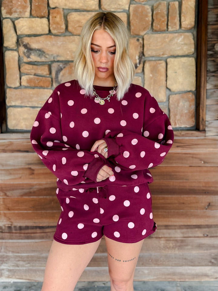 A woman with blonde hair stands on wooden steps in front of a stone wall, wearing a matching burgundy and white polka dot lounge set consisting of a sweatshirt and shorts.
