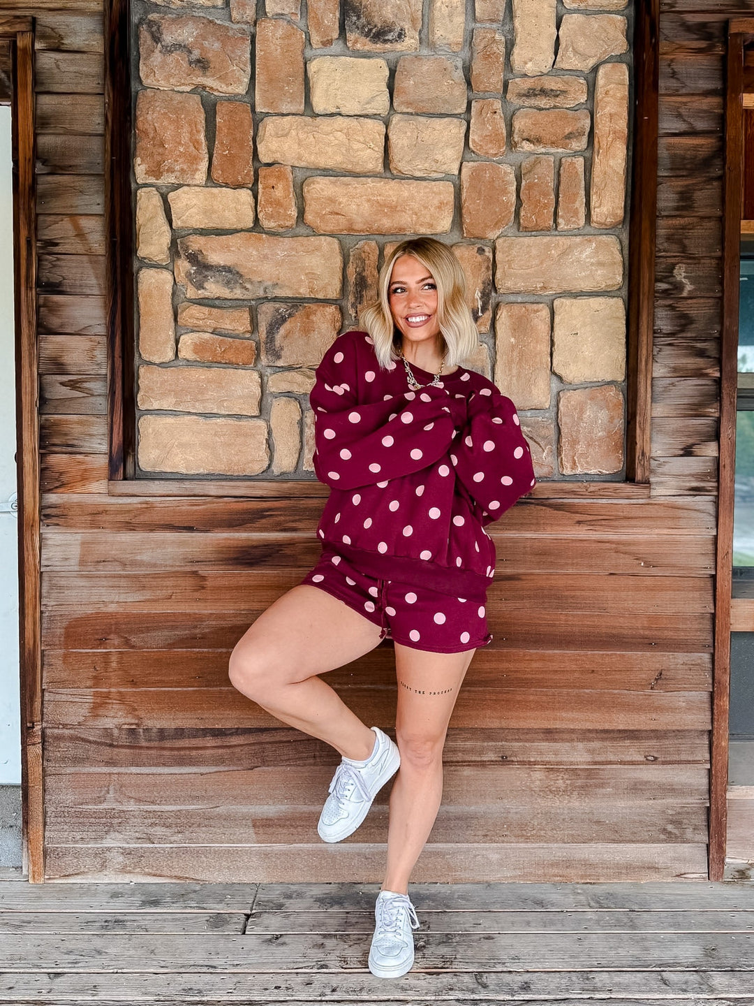 A woman posing in a matching burgundy and white polka dot lounge set, consisting of a long-sleeve sweatshirt and shorts, against a rustic stone and wood wall background.
