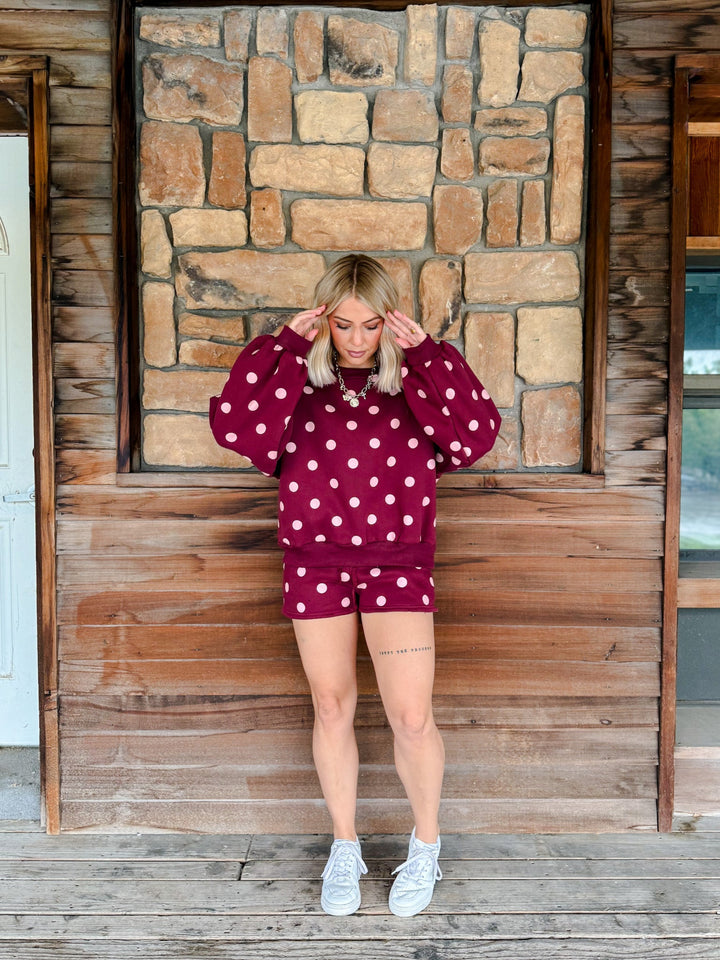 A woman stands on a wooden porch in front of a stone wall, wearing a matching burgundy and white polka dot lounge set consisting of a sweatshirt and shorts, paired with white sneakers.