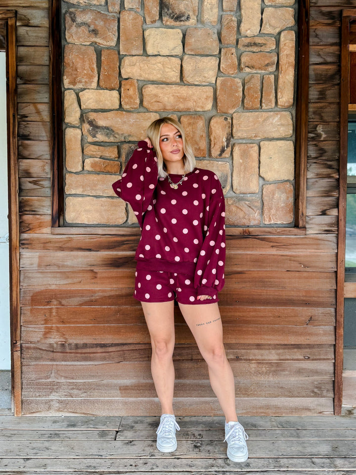 A woman stands in front of a rustic stone and wood wall wearing a matching burgundy and white polka dot lounge set consisting of a long-sleeve top and shorts, paired with white sneakers.