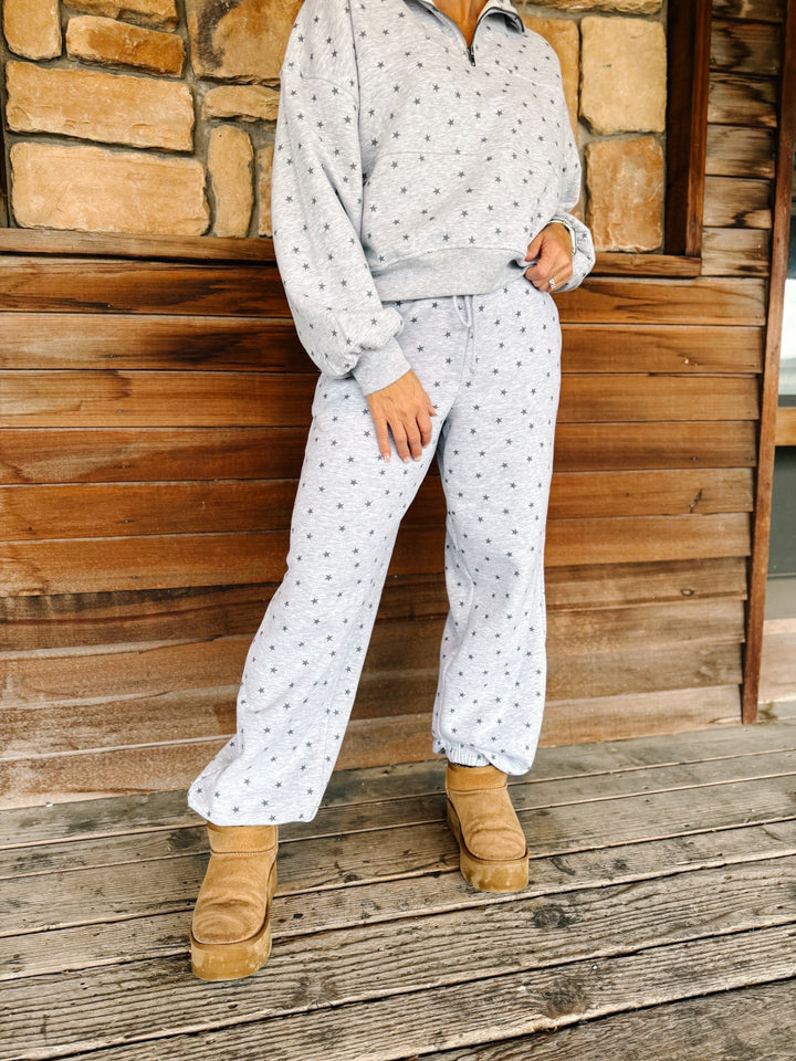 A person wearing a light gray and white star patterned sweatshirt and matching sweatpants, paired with tan suede boots, standing on a rustic wooden porch in front of a stone wall.