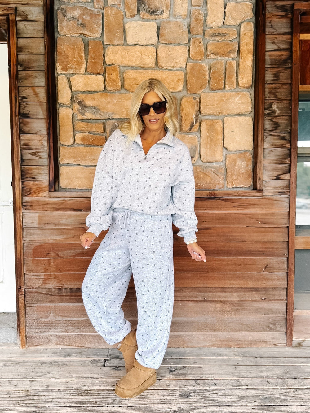 A person wearing a light gray and white star patterned sweatshirt and matching sweatpants, paired with tan suede boots, standing on a rustic wooden porch in front of a stone wall.