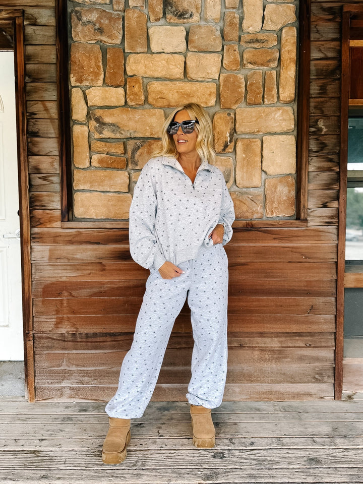 A person wearing a light gray and white star patterned sweatshirt and matching sweatpants, paired with tan suede boots, standing on a rustic wooden porch in front of a stone wall.