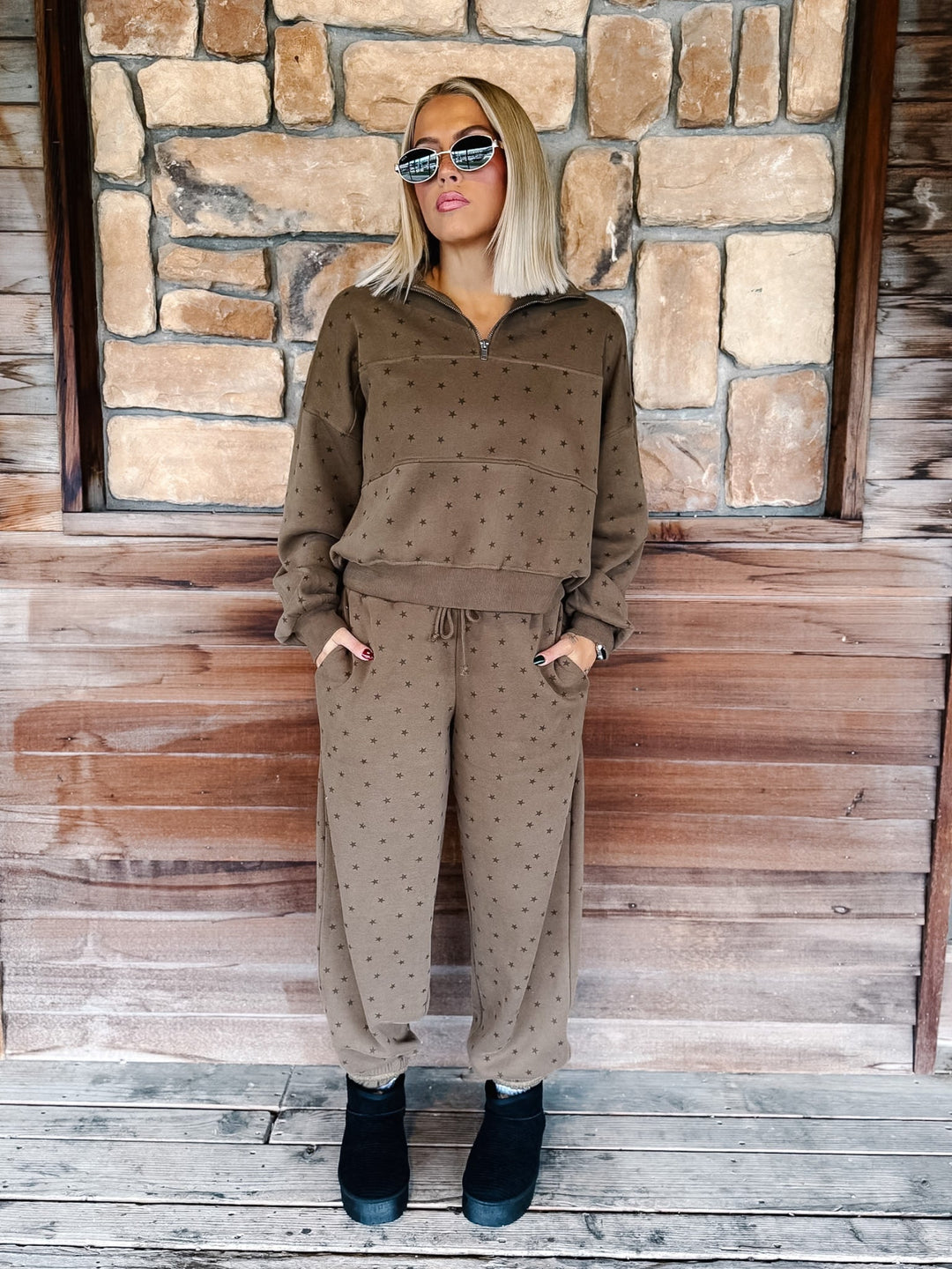 A woman stands on a wooden porch in front of a stone wall, wearing a matching brown half-zip sweatshirt and jogger pants set with a small star pattern, paired with black ankle boots.