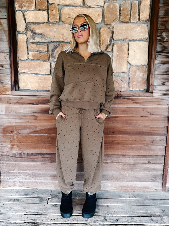 A woman stands on a wooden porch in front of a stone wall, wearing a matching brown half-zip sweatshirt and jogger pants set with a small star pattern, paired with black ankle boots.
