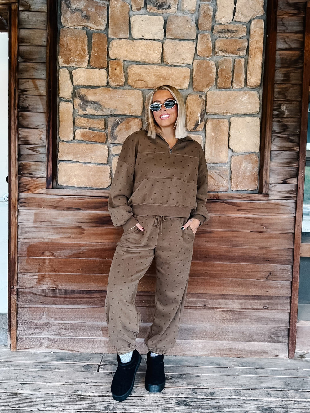 A woman stands on a wooden porch in front of a stone wall, wearing a matching brown half-zip sweatshirt and jogger pants set with a small star pattern, paired with black ankle boots.