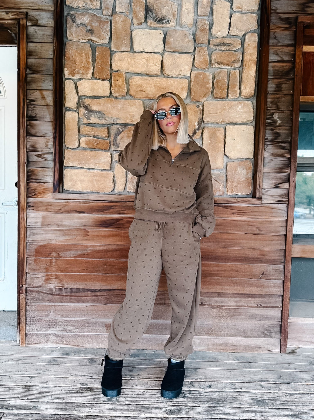A woman stands on a wooden porch in front of a stone wall, wearing a matching brown half-zip sweatshirt and jogger pants set with a small star pattern, paired with black ankle boots.