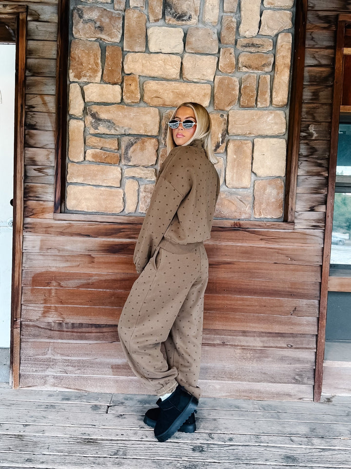 A woman stands on a wooden porch in front of a stone wall, wearing a matching brown half-zip sweatshirt and jogger pants set with a small star pattern, paired with black ankle boots.