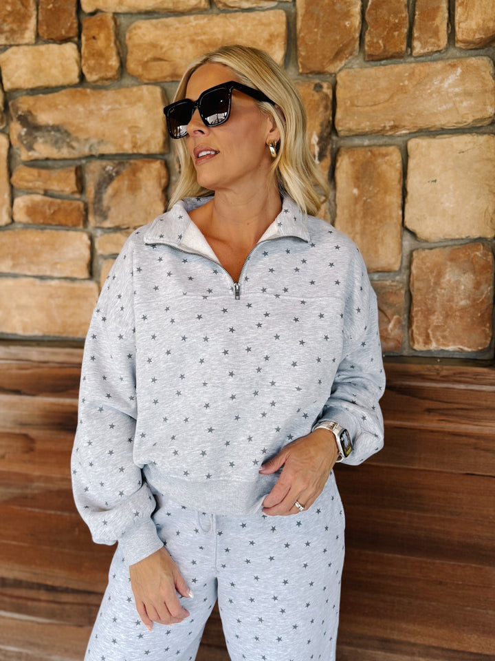 A woman wearing a light gray heathered quarter-zip sweatshirt and matching sweatpants with a small dark star pattern, standing in front of a rustic stone wall.
