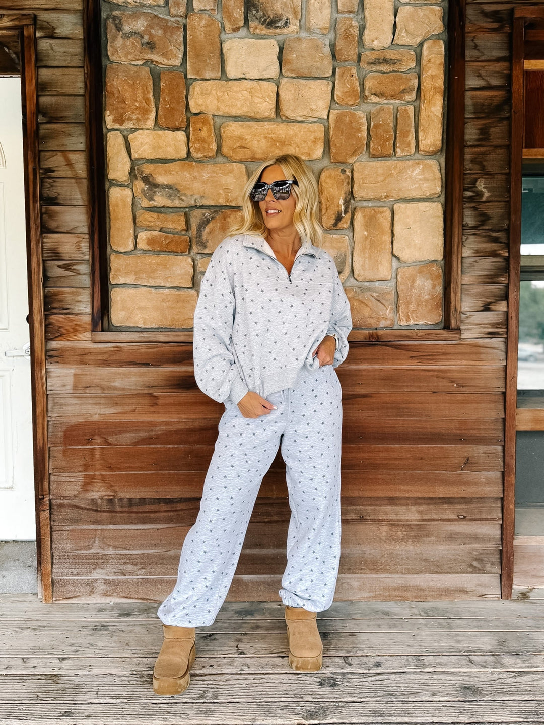 A woman wearing a light heather grey  star patterned matching lounge set stands on a wooden porch in front of a stone wall