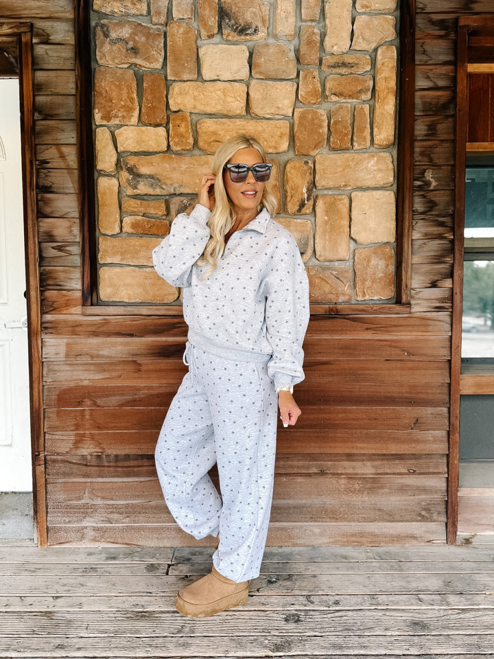 A woman wearing a light gray star patterned matching lounge set stands on a rustic wooden porch in front of a stone wall.