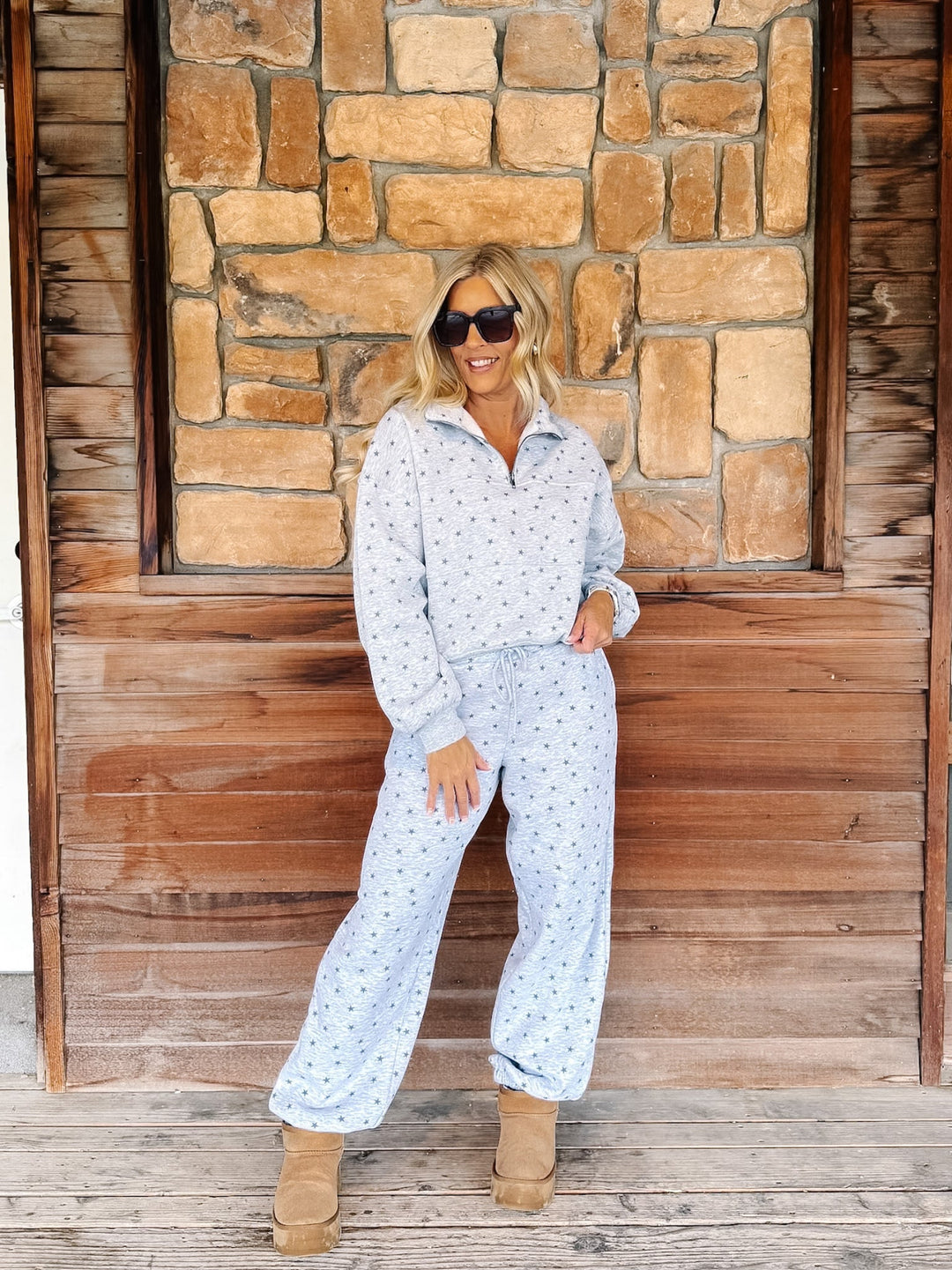 A woman wearing a light grey star patterned matching lounge set stands in front of a rustic stone and wood wall, wearing tan boots and sunglasses.