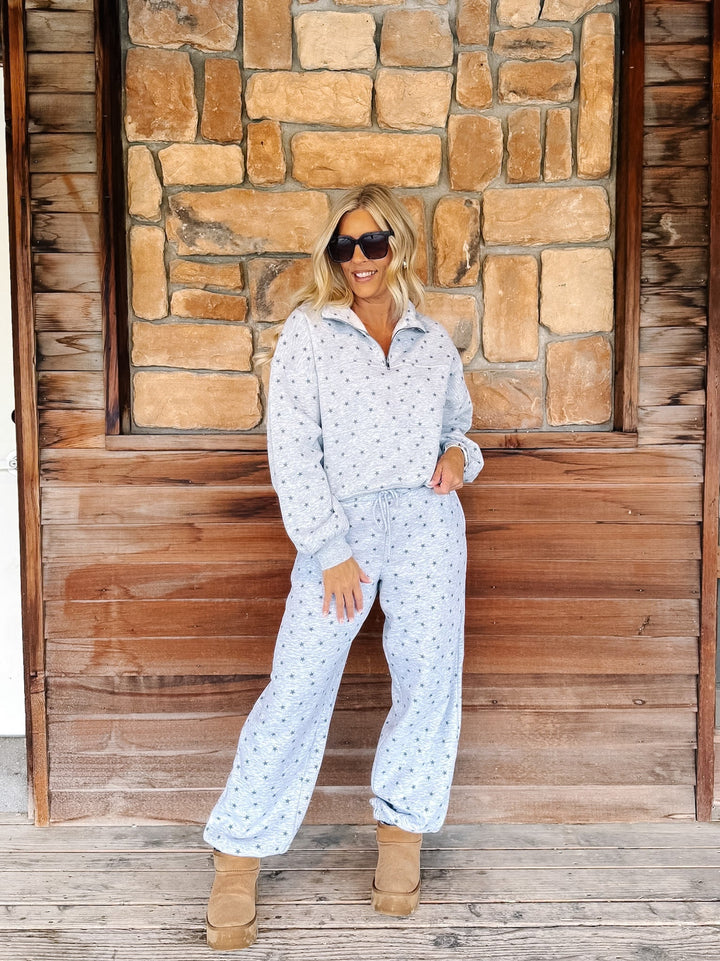 A woman wearing a light grey star patterned matching lounge set stands in front of a rustic stone and wood wall, wearing tan boots and sunglasses.