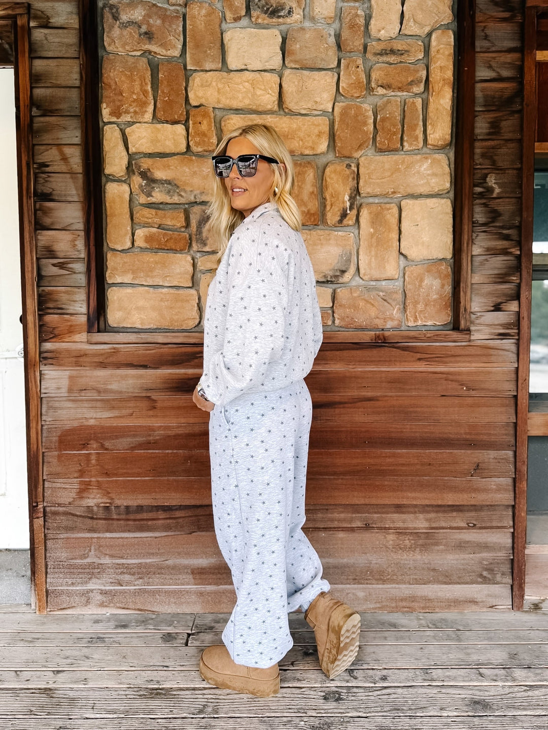 A woman wearing a heather grey star top with matching joggers, styled with tan boots, standing in front of a rustic stone and wood wall.