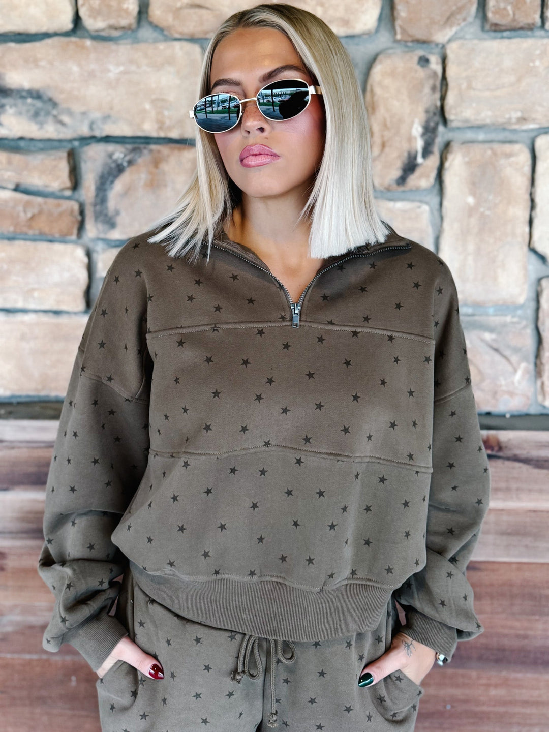 A woman with blonde hair and sunglasses wearing an mocha brown star-patterned half-zip sweatshirt and matching sweatpants, standing in front of a stone wall.