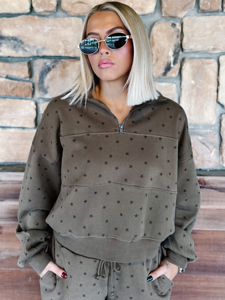 A woman with blonde hair and sunglasses wearing an mocha brown star-patterned half-zip sweatshirt and matching sweatpants, standing in front of a stone wall.