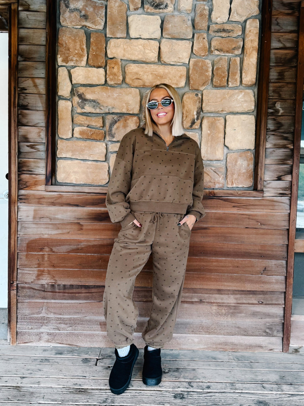 A woman with blonde hair and sunglasses wearing an mocha brown star-patterned half-zip sweatshirt and matching sweatpants, standing in front of a stone wall.