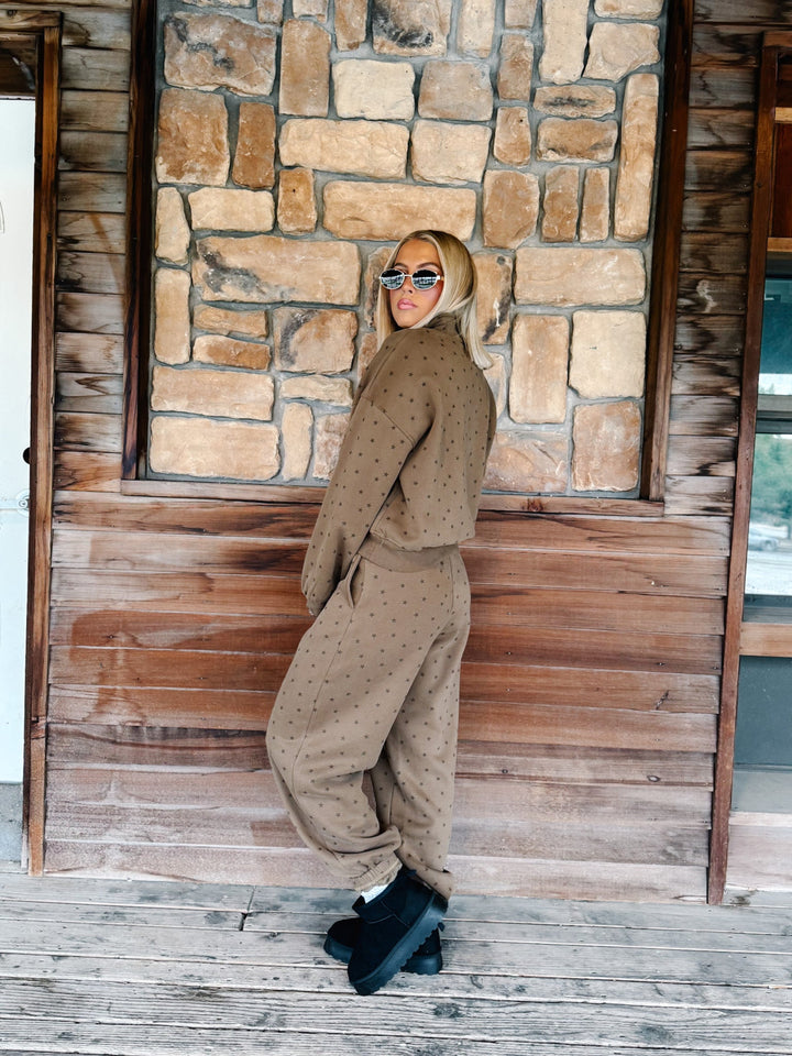 A woman with blonde hair and sunglasses wearing an mocha brown star-patterned half-zip sweatshirt and matching sweatpants, standing in front of a stone wall.