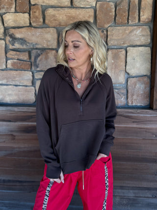 A woman wearing a dark brown half-zip hoodie and bright pink track pants with a black-and-white patterned side stripe, standing in front of a rustic stone wall.