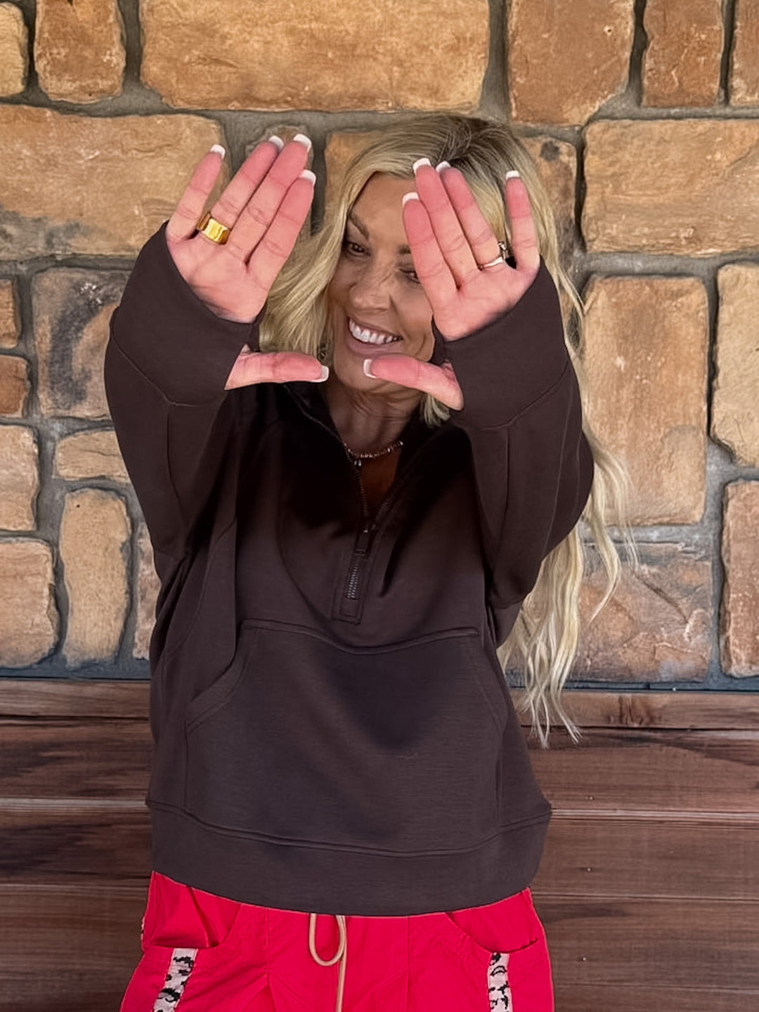 A woman with blonde hair wearing a dark brown half-zip sweatshirt and bright red sweatpants, posing with her hands in front of her face against a rustic stone wall background.