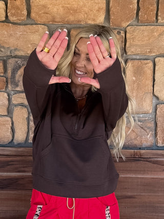 A woman with blonde hair wearing a dark brown half-zip sweatshirt and bright red sweatpants, posing with her hands in front of her face against a rustic stone wall background.