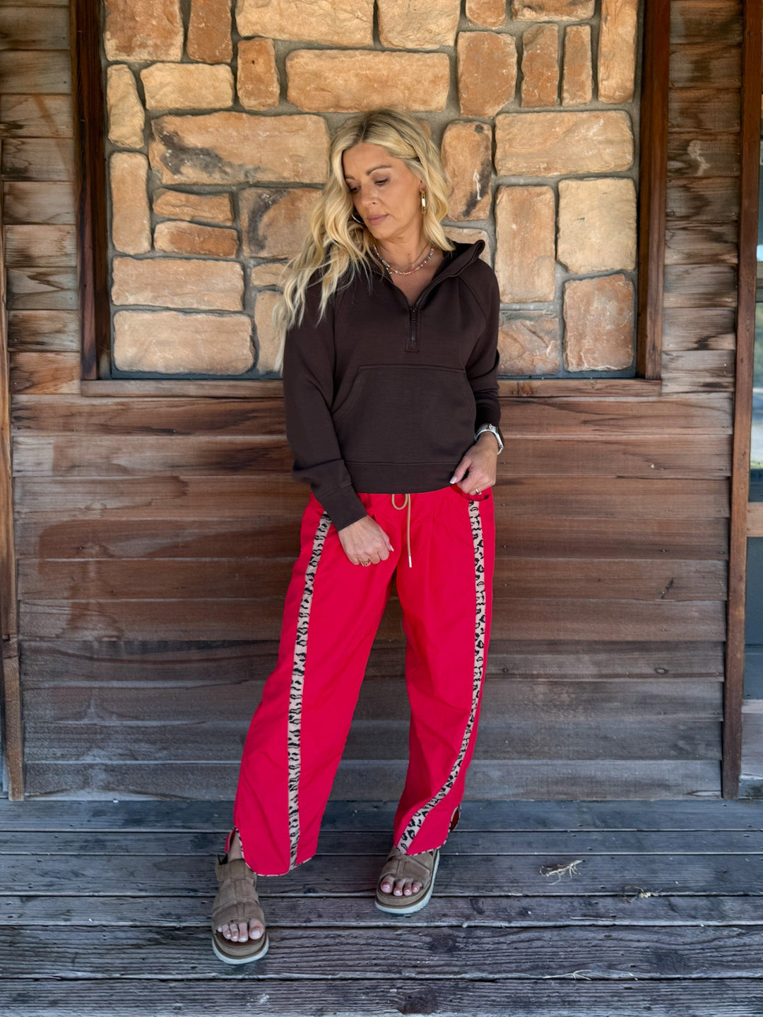 A woman stands on a wooden porch in front of a stone wall, wearing bright red drawstring sweatpants with white chain-like side stripes, a dark brown long-sleeve top, and brown sandals.