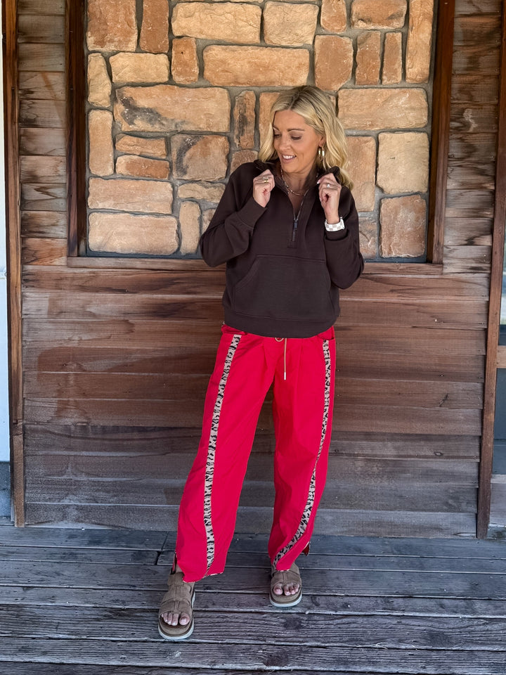A woman stands on a rustic wooden porch in front of a stone wall, wearing a dark brown half-zip pullover and bright red sweatpants with white text down the sides.
