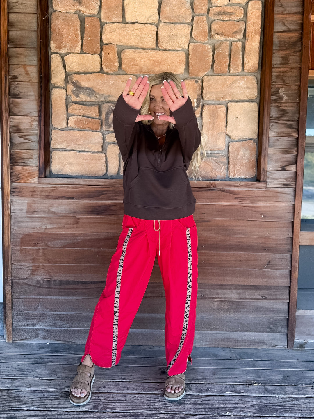 A person standing on a wooden deck in front of a stone wall, wearing a dark brown hoodie and bright red sweatpants with a patterned side stripe, holding their hands up in front of their face.