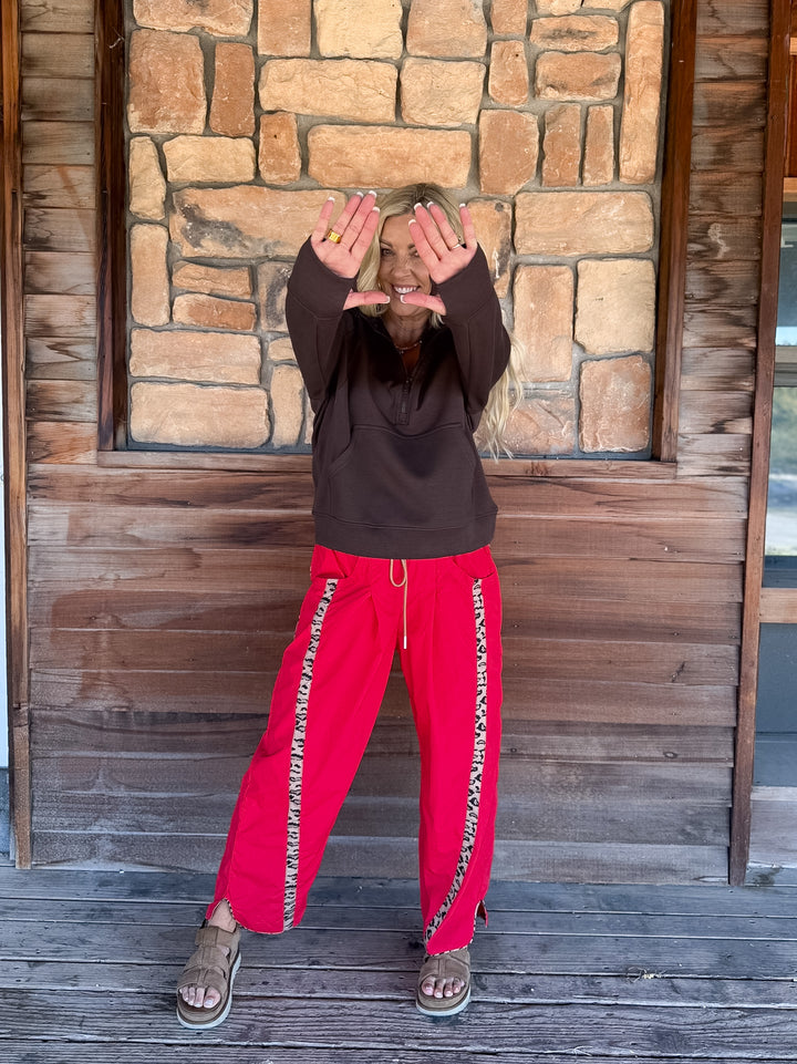 A person standing on a wooden deck in front of a stone wall, wearing a dark brown hoodie and bright red sweatpants with a patterned side stripe, holding their hands up in front of their face.