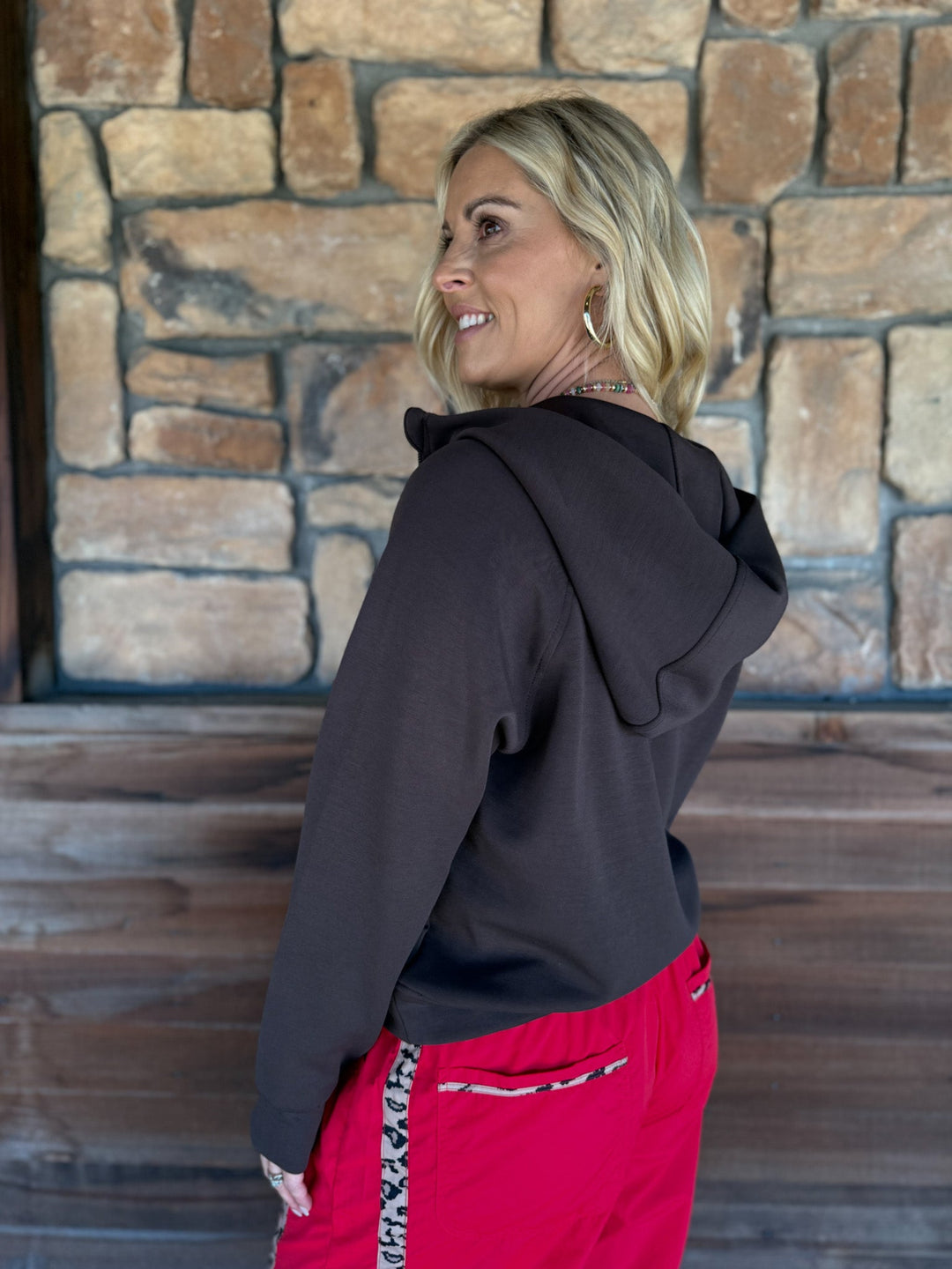 A woman with blonde hair is shown from the back wearing a dark brownish-black hoodie and bright pink pants with a leopard print side stripe, standing in front of a rustic stone wall.