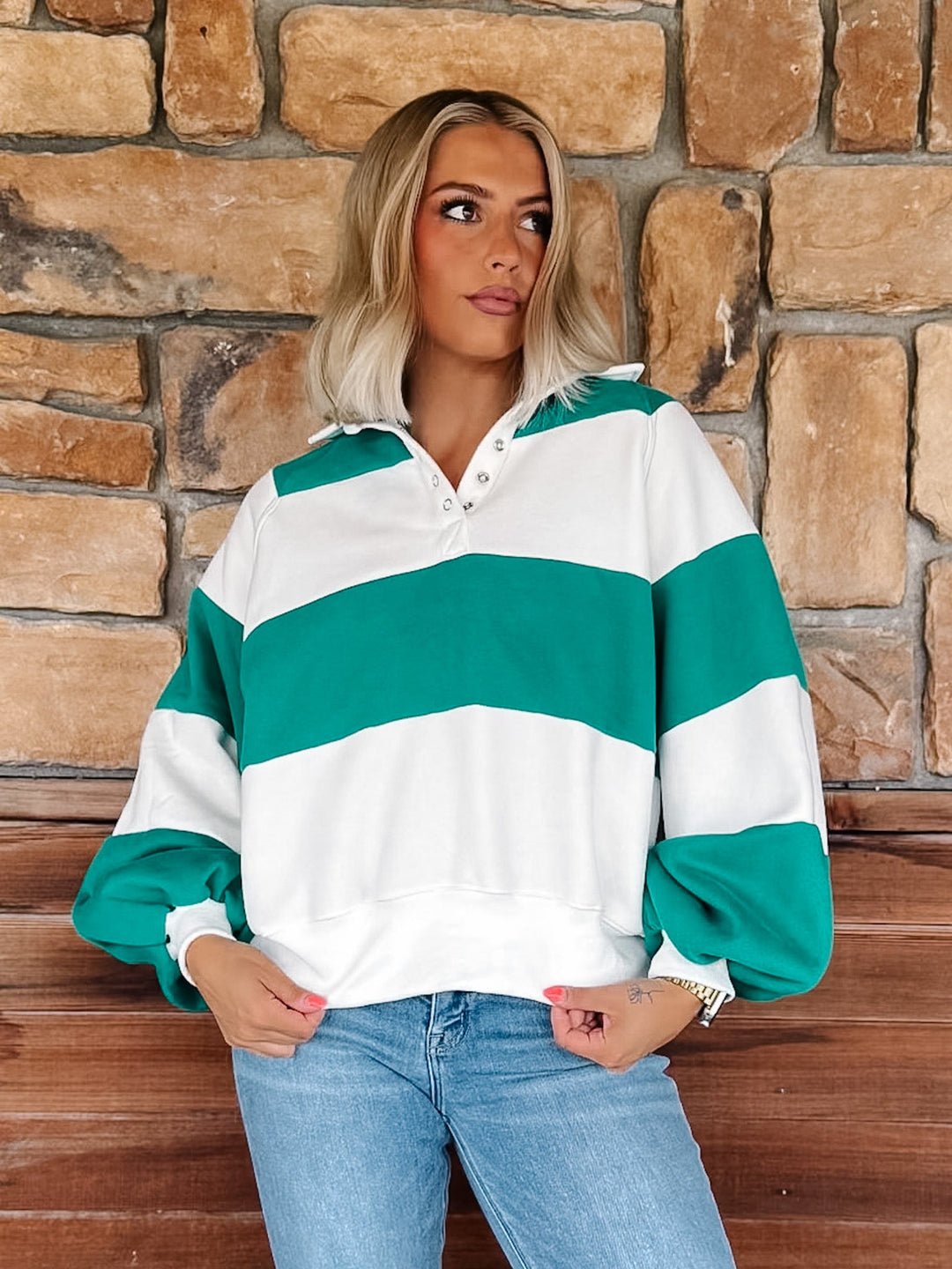 A woman wearing a green and white striped long-sleeve rugby pullover sweatshirt with a half-placket collar, paired with light-wash blue jeans, standing in front of a rustic stone wall.