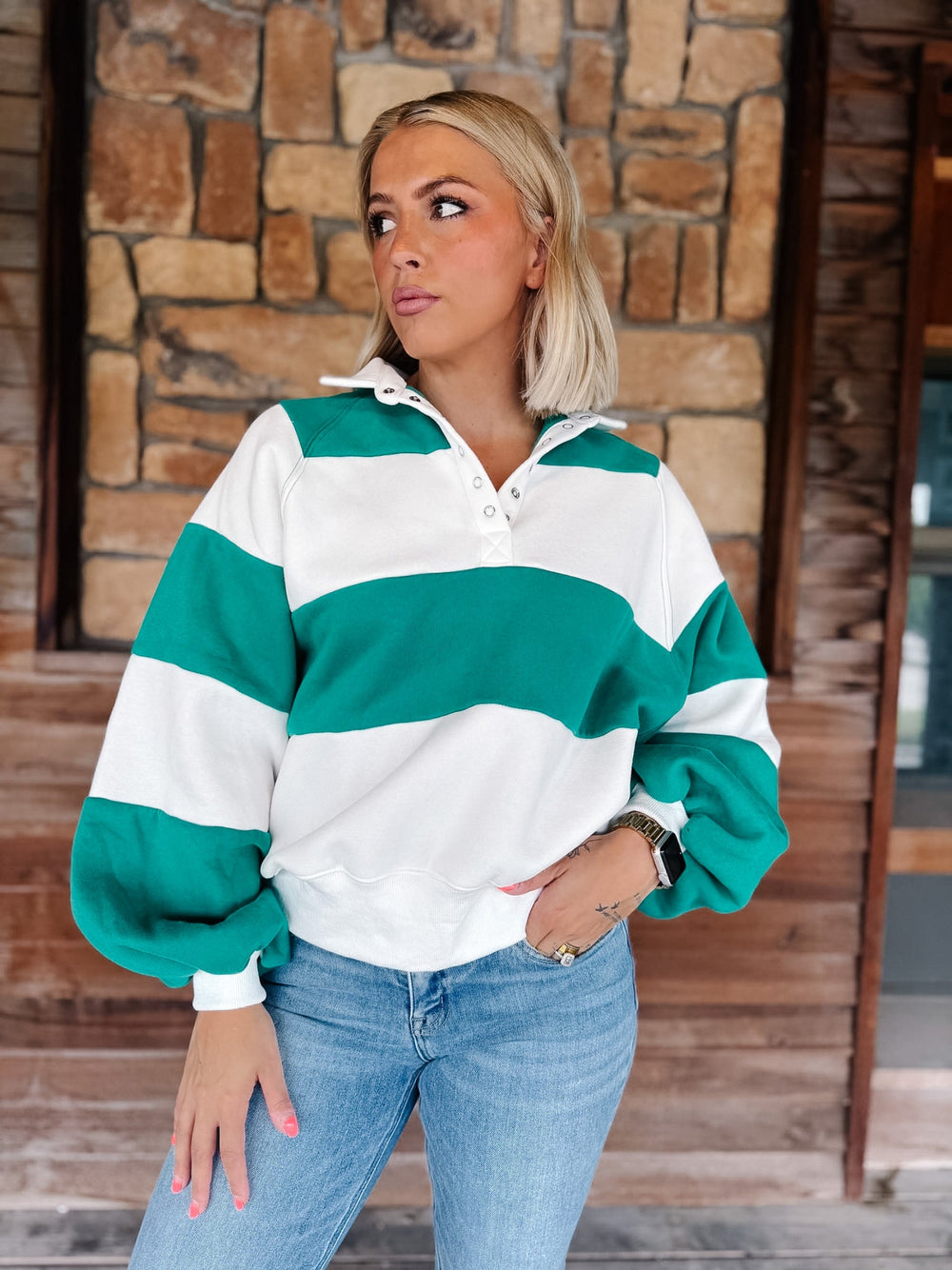 A woman wearing a green and white horizontally striped long-sleeve rugby-style pullover sweater with a partial button placket, paired with light blue jeans, standing in front of a rustic stone and wood wall.