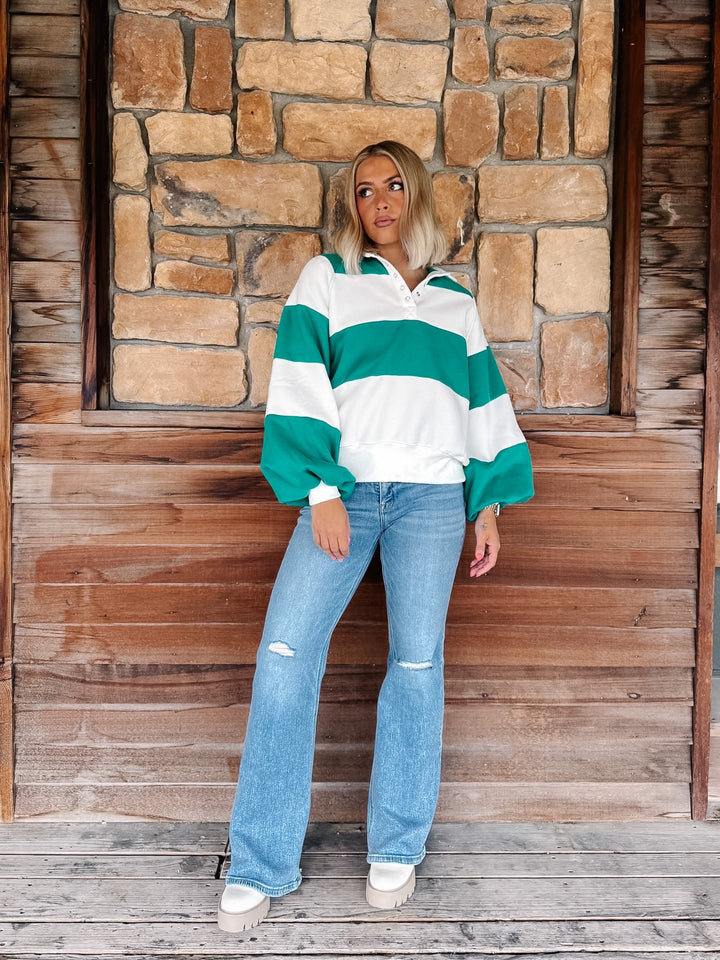 A woman stands on a wooden porch in front of a stone wall, wearing a green and white striped long-sleeve pullover sweatshirt with balloon sleeves, light-wash distressed jeans, and white sneakers.