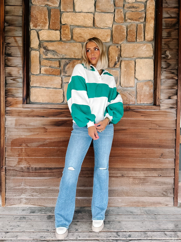 A woman stands on a wooden porch in front of a stone wall, wearing a green and white horizontally striped long-sleeve sweatshirt, light blue distressed jeans, and white sneakers.
