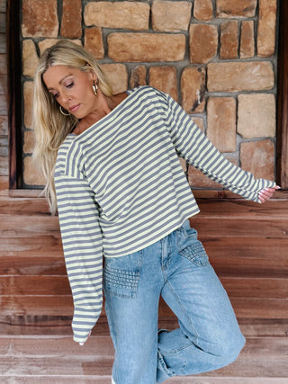 A woman wearing a boat neck striped top in grey and yellow, paired with light blue jeans, posing in front of a rustic stone wall.