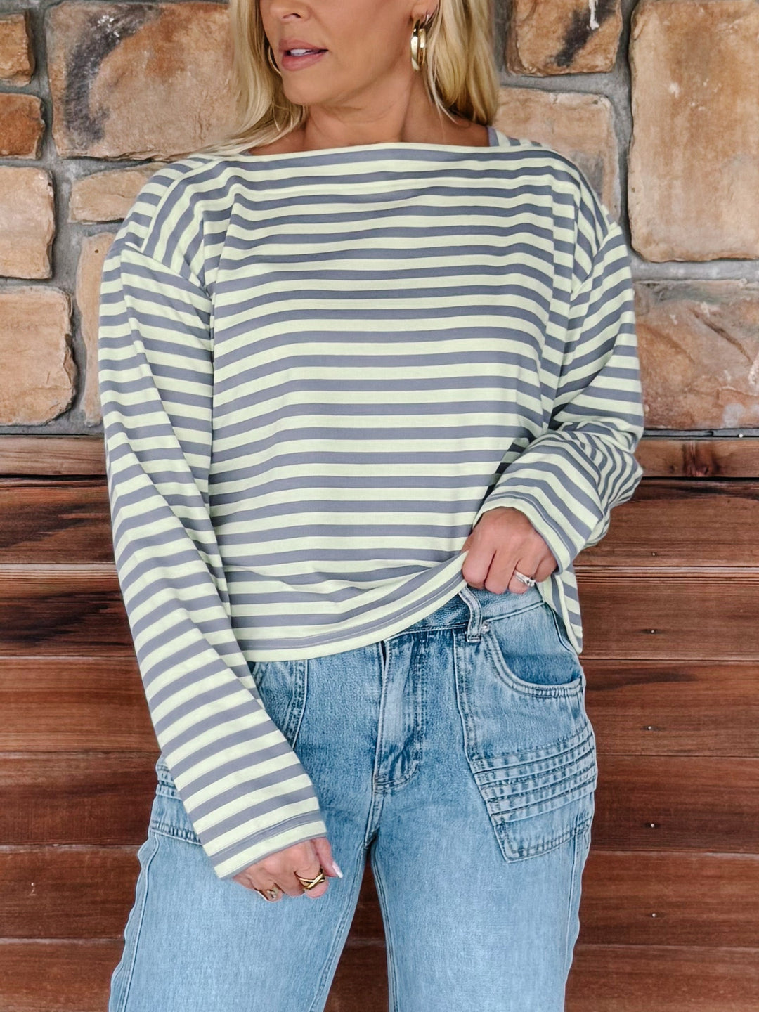 A woman wearing a boat neck striped top in grey and yellow, paired with light blue jeans, posing in front of a rustic stone wall.