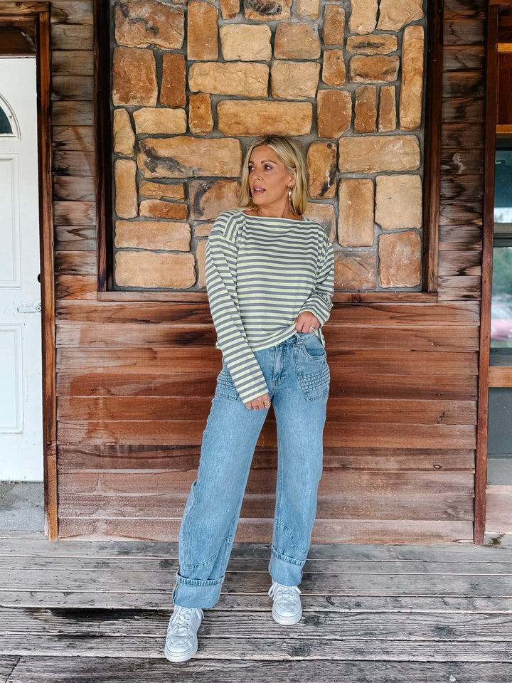 A woman wearing a boat neck striped top in grey and yellow, paired with light blue jeans, posing in front of a rustic stone wall.