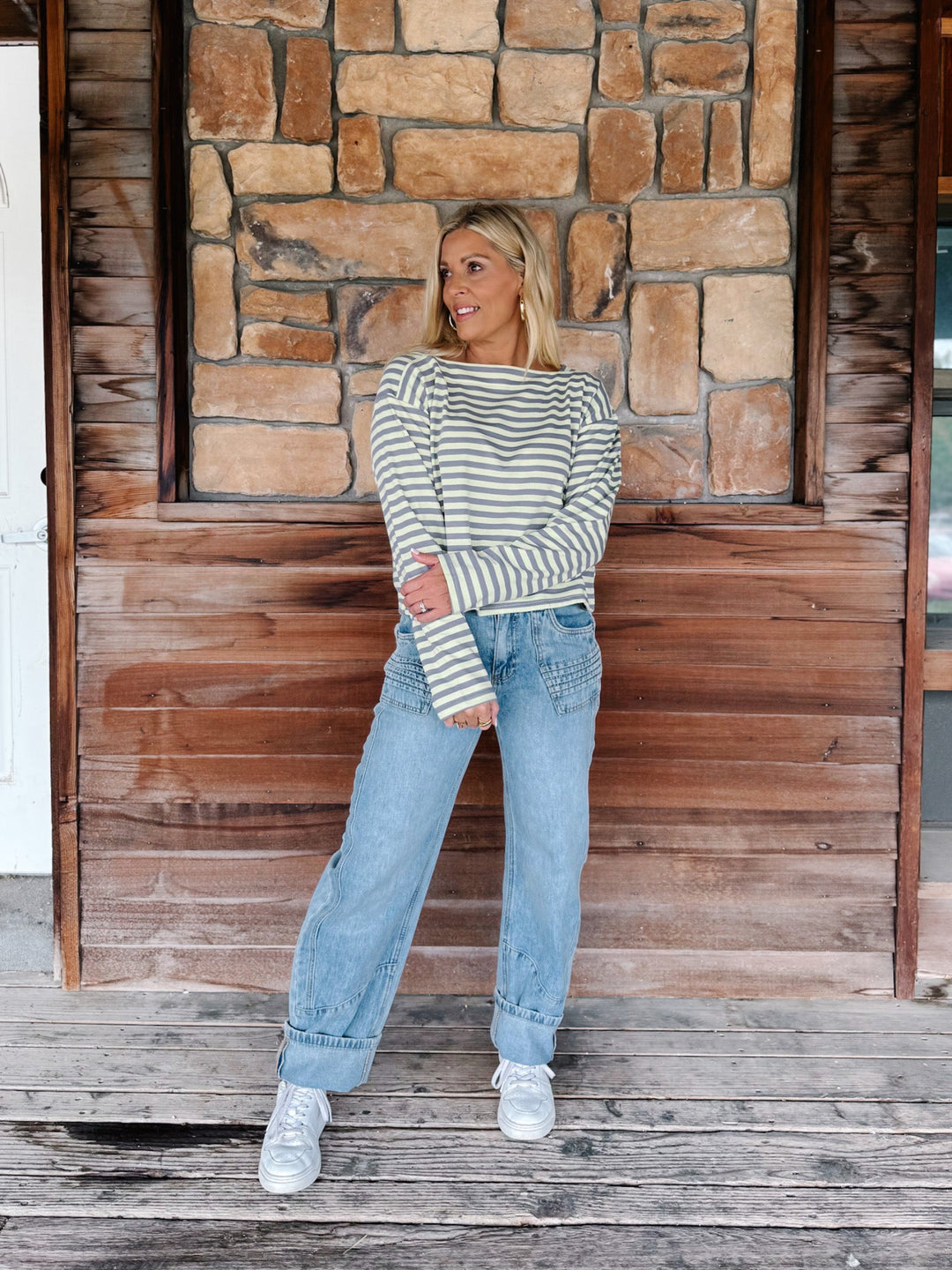 A woman wearing a boat neck striped top in grey and yellow, paired with light blue jeans, posing in front of a rustic stone wall.