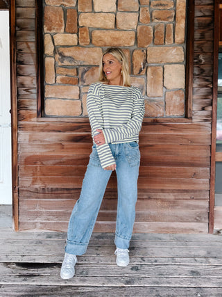 A woman wearing a boat neck striped top in grey and yellow, paired with light blue jeans, posing in front of a rustic stone wall.