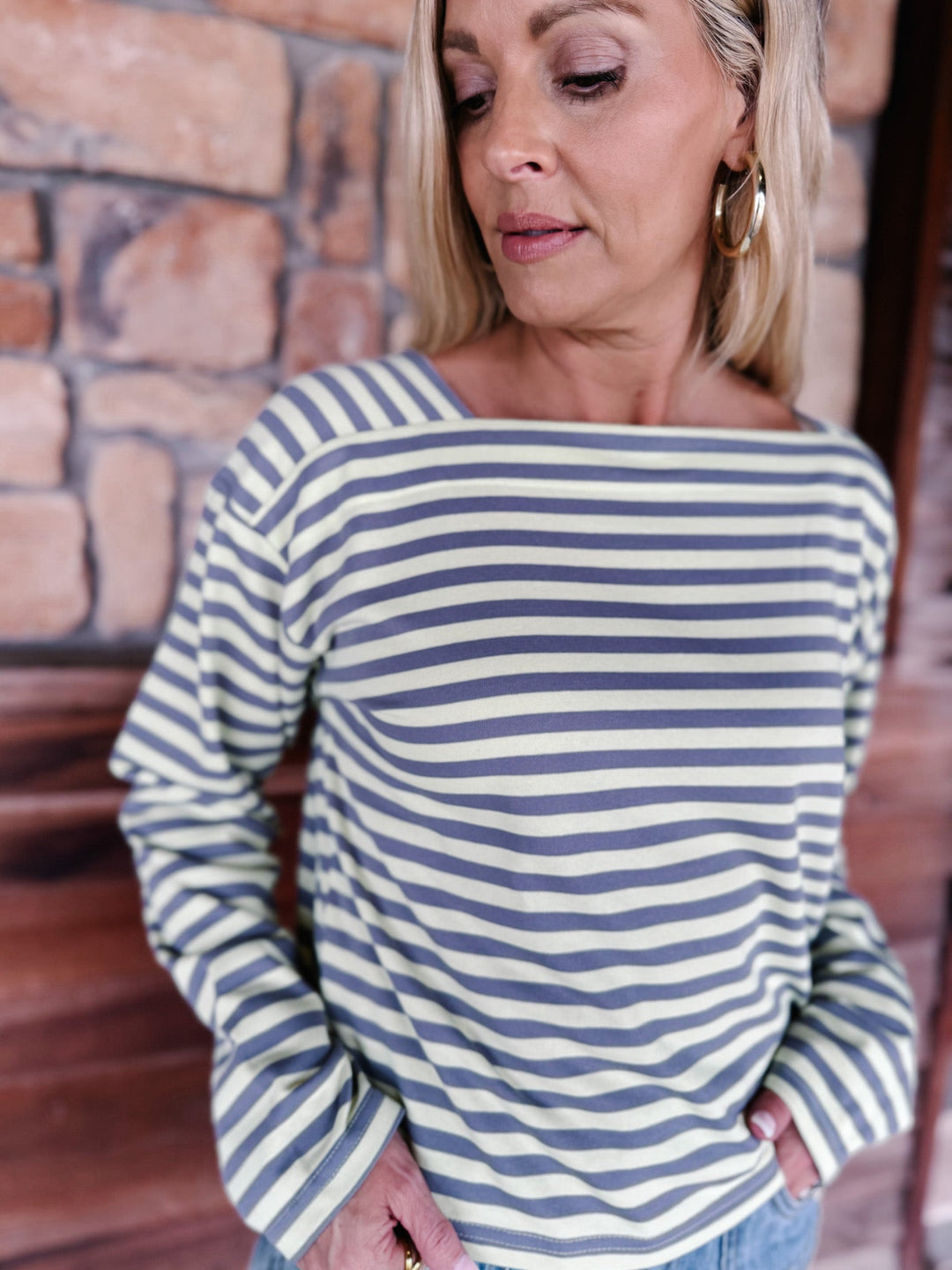 A woman wearing a boat neck striped top in grey and yellow, paired with light blue jeans, posing in front of a rustic stone wall.