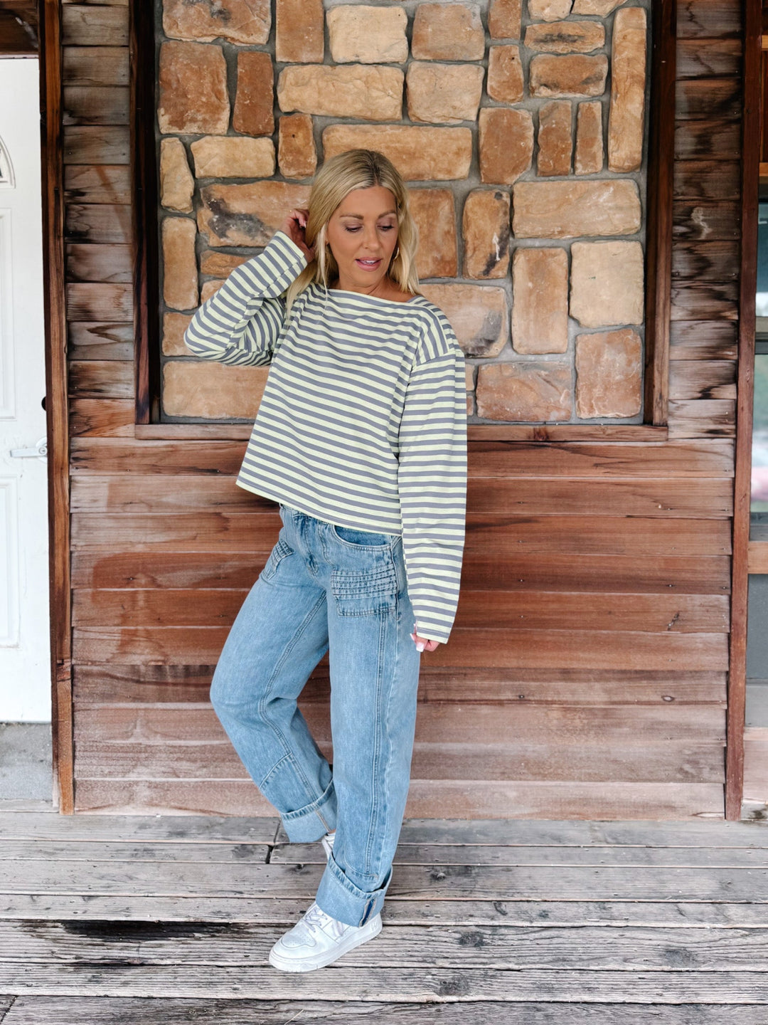A woman wearing a boat neck striped top in grey and yellow, paired with light blue jeans, posing in front of a rustic stone wall.