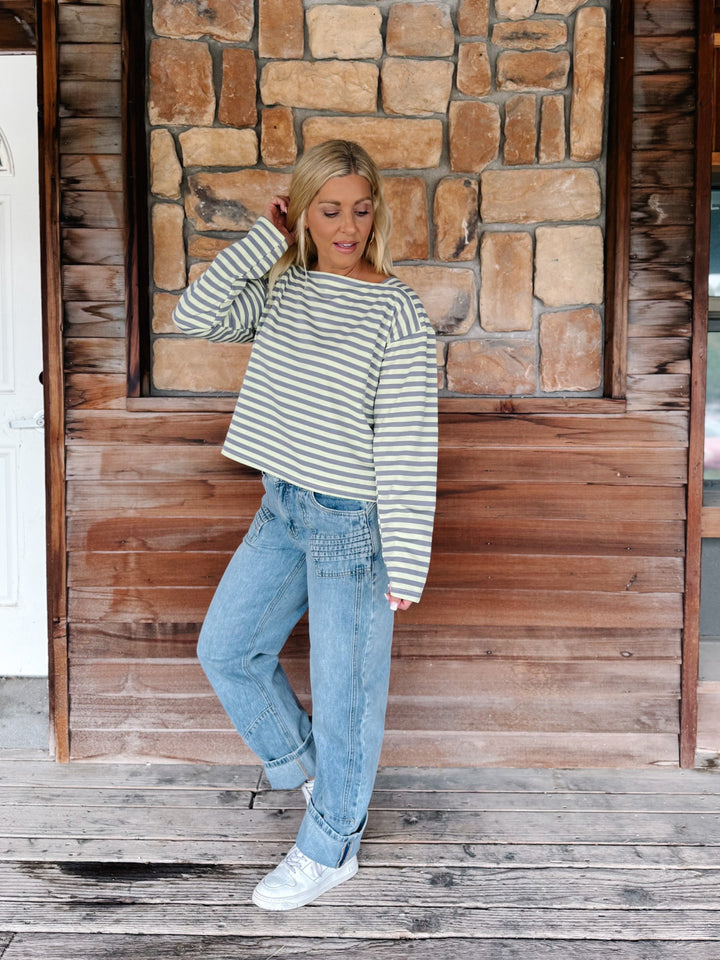 A woman wearing a boat neck striped top in grey and yellow, paired with light blue jeans, posing in front of a rustic stone wall.