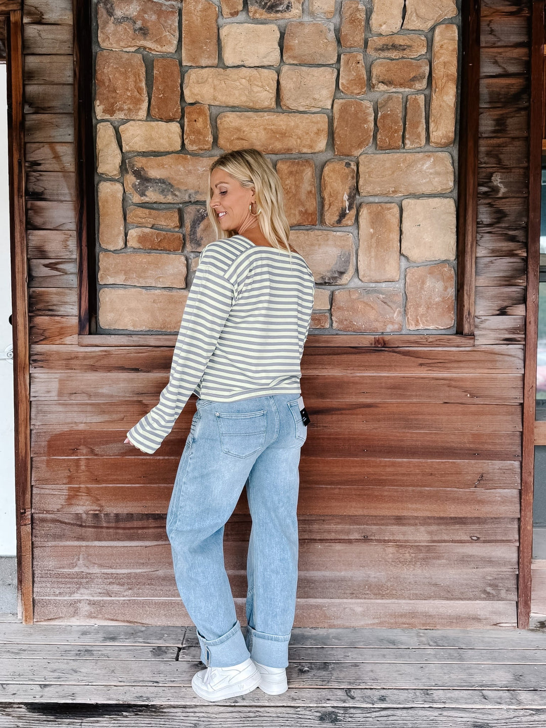 A woman wearing a boat neck striped top in grey and yellow, paired with light blue jeans, posing in front of a rustic stone wall.