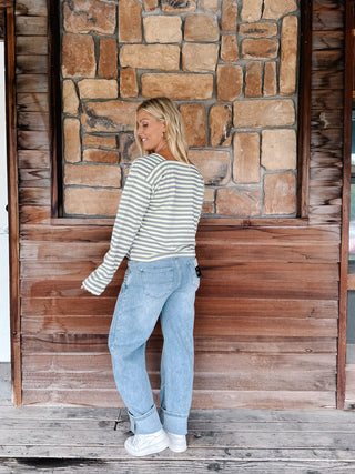 A woman wearing a boat neck striped top in grey and yellow, paired with light blue jeans, posing in front of a rustic stone wall.